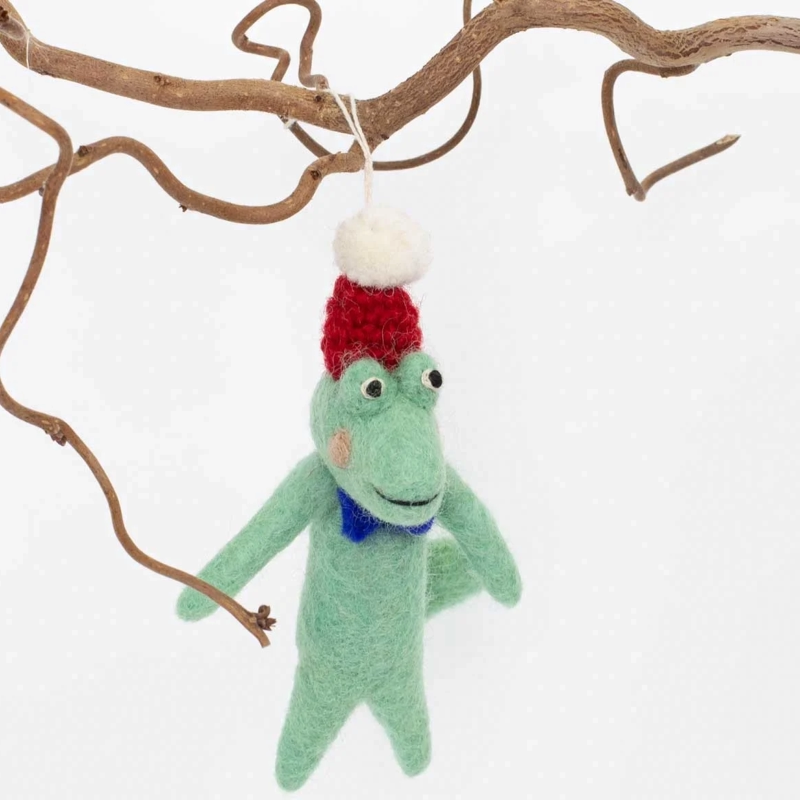 Julgranshänge i form av en tovad grön ullkrokodil med röd mössa och blå fluga som hänger på en gren.