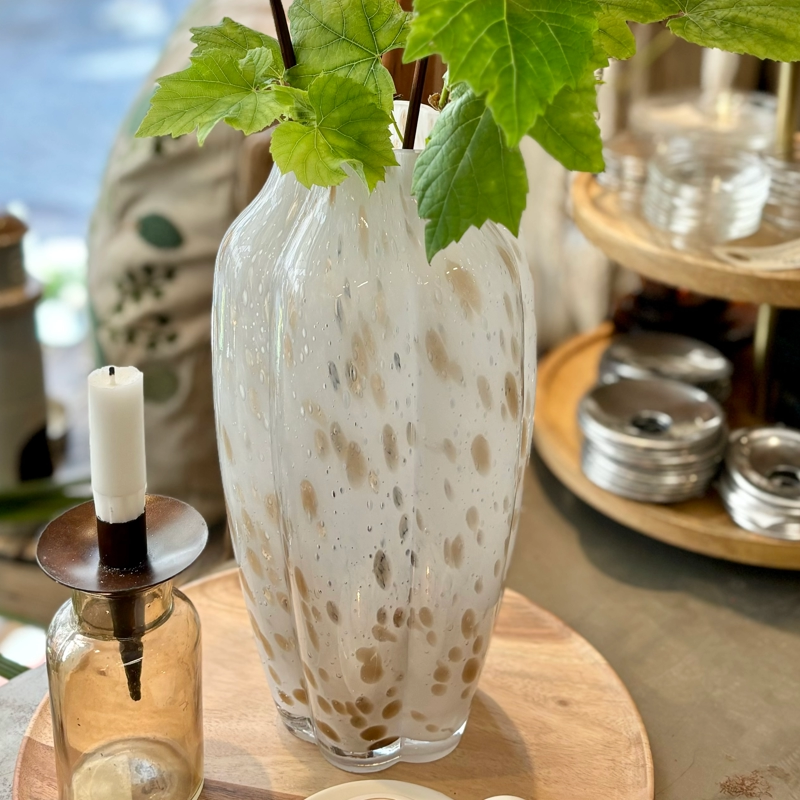 Hög vit glasvas med beige stänk på, ståendes på en rund träbricka.